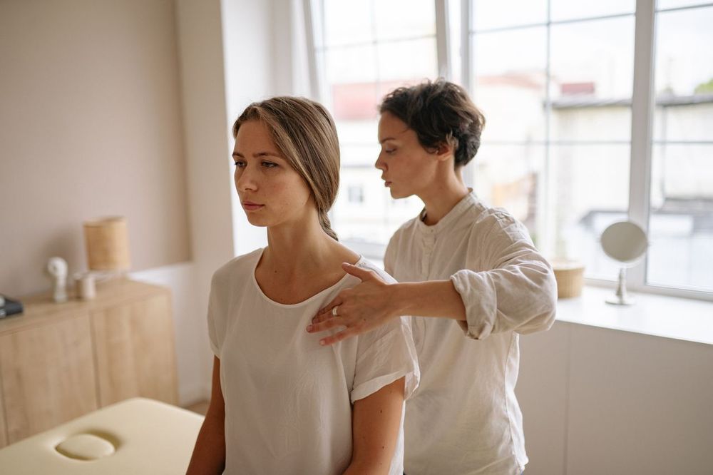 Zwei Frauen in einem hellen Raum am Fenster: Eine Lehrerin legt von hinten behutsam die Hand an die Schulter einer aufrecht stehenden Frau